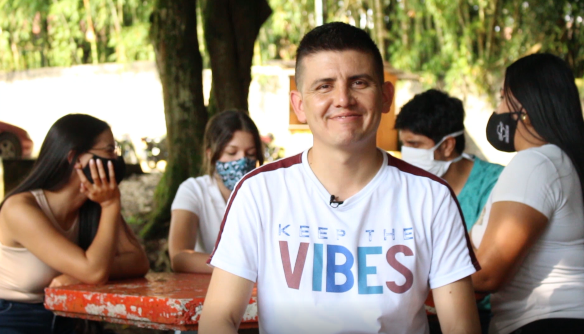 Estudiante sonriendo frente a la cámara, mientras un grupo de estudiantes hablan y comparten en segundo plano. 