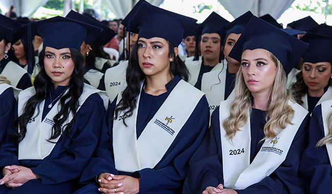 Tres estudiantes con toga y birrete, prestando atención a la ceremonia de graduación. 