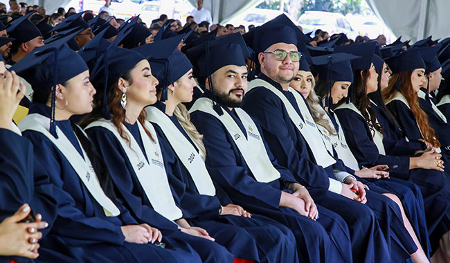 Estudiantes sentados con toga y birrete, prestando atención a la ceremonia de graduación. 