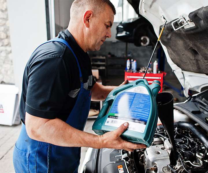 Esta imagen muestra a un mecánico realizando el cambio de aceite de un automóvil en un taller. Es un hombre de mediana edad con el cabello corto y canoso, vestido con un uniforme compuesto por una camiseta negra y un delantal azul