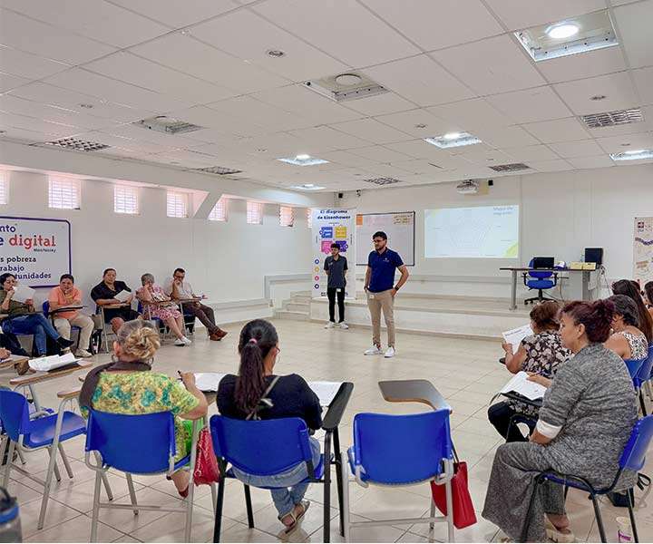 En un aula amplia e iluminada, un hombre con camisa azul y pantalón beige se encuentra de pie en el centro, hablando con los asistentes. A su lado, otro hombre con ropa oscura observa. Alrededor, varias personas, en su mayoría mujeres, están sentadas en sillas azules con escritorios adjuntos, tomando notas y prestando atención. En el fondo, hay una pantalla proyectando información, indicando un entorno educativo
