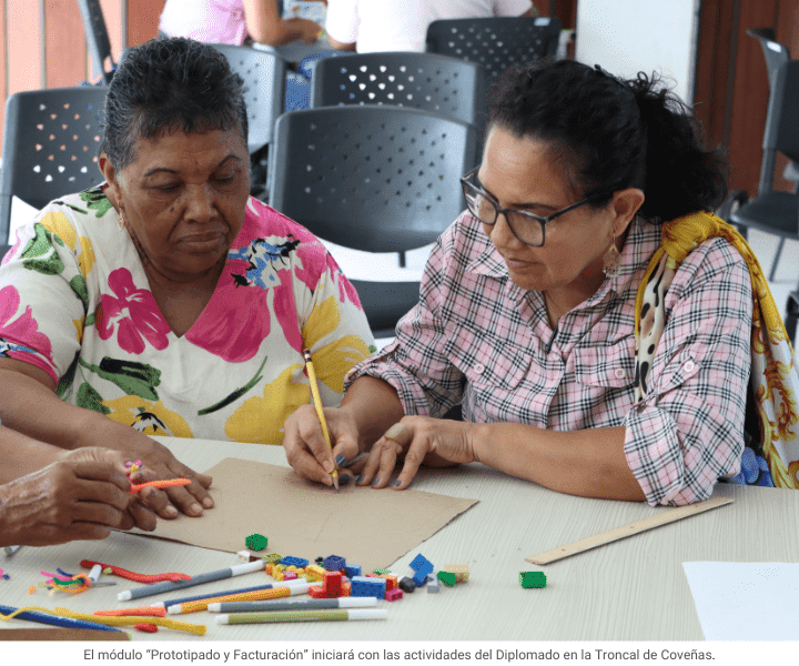 Dos mujeres mayores trabajan juntas en una actividad grupal. Una, con blusa floreada, observa atentamente mientras la otra, con camisa a cuadros y gafas, dibuja sobre un cartón. En la mesa hay lápices, plastilina y bloques de colores.