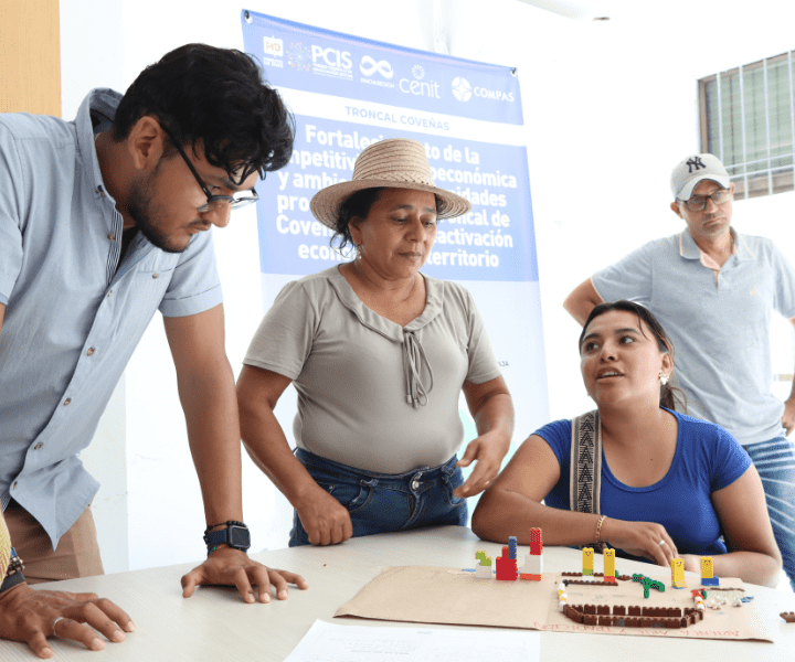 Cuatro personas participan en una actividad grupal en un aula iluminada. Una mujer con sombrero de paja habla mientras los demás la escuchan atentamente. En la mesa hay una maqueta con bloques de colores. Al fondo, un cartel menciona el fortalecimiento económico en la Troncal de Coveñas.