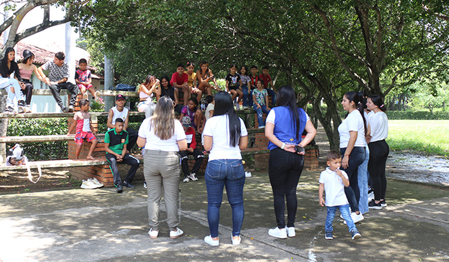 Un grupo de personas, incluidos niños, participan en una actividad grupal al aire libre, dirigida por docentes y estudiantes de Trabajo Social