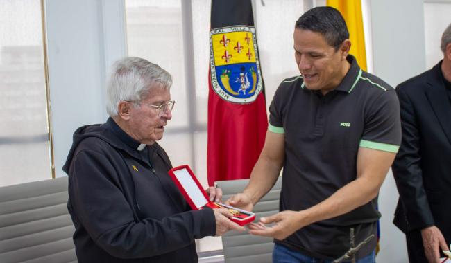Alcalde de Cúcuta, Jorge Acevedo, entrega una condecoración al Padre Diego Jaramillo durante un acto oficial. Al fondo, la bandera de la ciudad y símbolos institucionales.