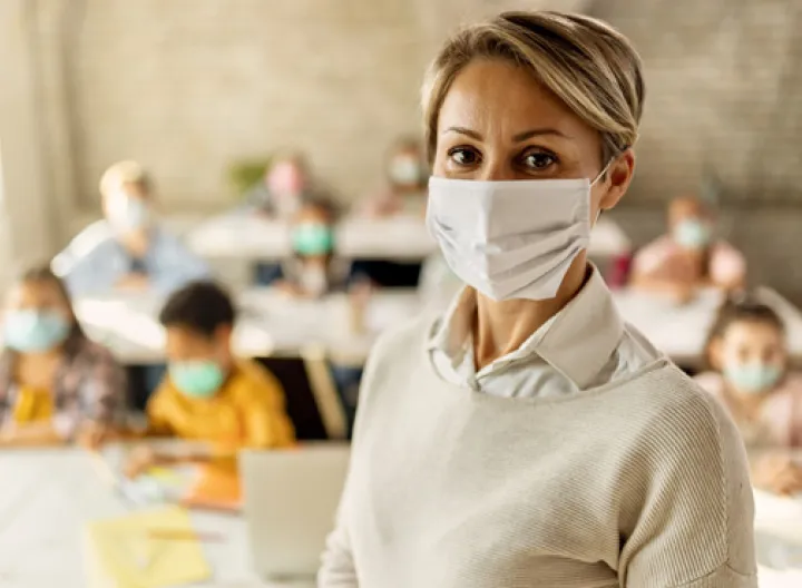 Mujer con tapabocas en salón de clases, de fondo se ve un grupo de niños sentados en sus puestos