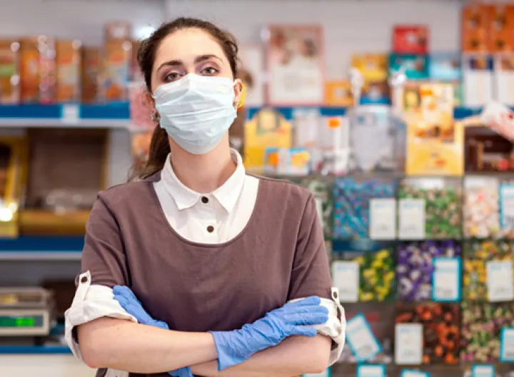 Mujer cruzada de brazos con tapabocas y guantes, de fondo se ve una papelería