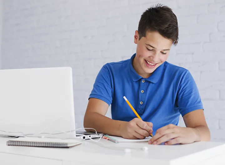 Un hombre joven camiseta azul sentado en un escritorio con un computador ey escribiendo en un cuaderno.