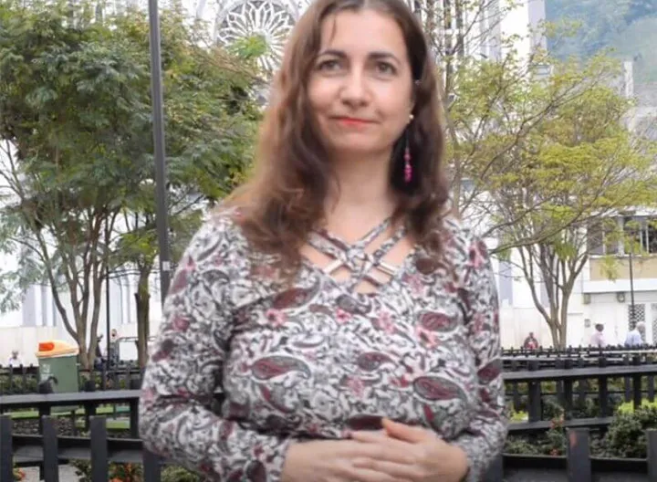 Docente en el centro de la imagen, con sus manos entrecruzadas en el abdomen, está de pie. Con una pequeña sonrisa y su cabello ondulado, mira al frente. Al fondo se ve el parque principal de chinchiná con unos arboles que dejan ver u poco de la fachada de la iglesia principa