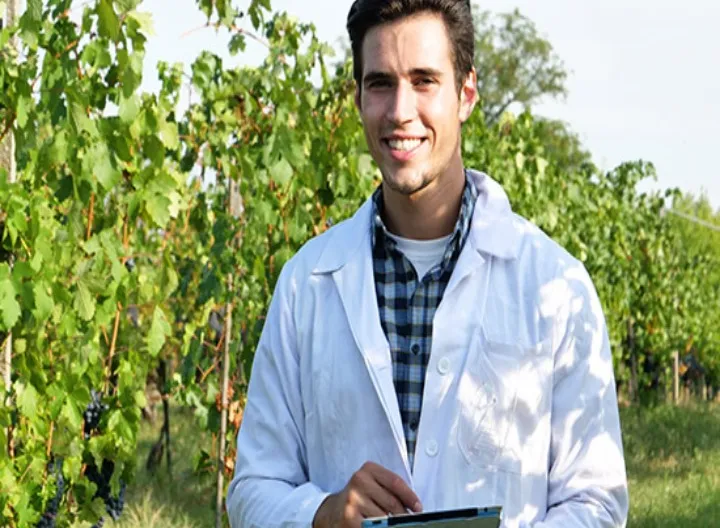 Mirando al frete: Joven con con sonrisa, viste una bata blanca y sostiene una tablet,mientras realiza labores de estudio en el campo. Al fondo un cultivo alto.