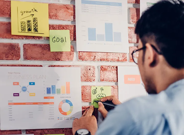 Hombre de espalda usando camisa azul y gafas organizado ideas en una cartelera
