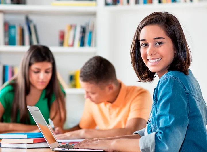 Joven estudiante en mesa de biblioteca, con su computador frente, mira a la cámara con el cuerpo en dirección al portatil y su rostro girado. Al fondo se ven dos jóvenes estudiando y dialogando. 