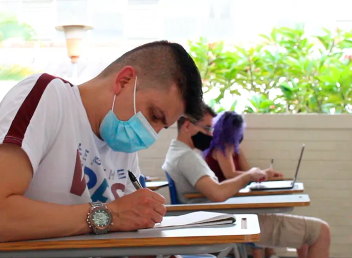 Estudiante usa tapaboca y escribe en silla de universidad. Al fondo se ven dos compañeros suños escuchando la clase. 