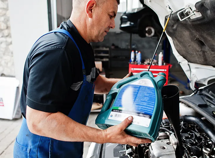 Esta imagen muestra a un mecánico realizando el cambio de aceite de un automóvil en un taller. Es un hombre de mediana edad con el cabello corto y canoso, vestido con un uniforme compuesto por una camiseta negra y un delantal azul