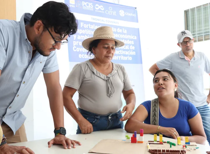 Cuatro personas participan en una actividad grupal en un aula iluminada. Una mujer con sombrero de paja habla mientras los demás la escuchan atentamente. En la mesa hay una maqueta con bloques de colores. Al fondo, un cartel menciona el fortalecimiento económico en la Troncal de Coveñas.
