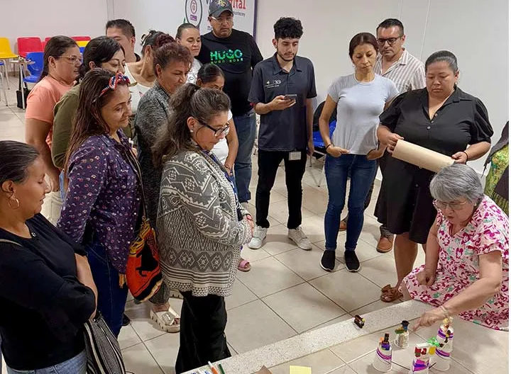 Varias personas están de pie alrededor de una mesa, con una mujer en el medio, creando un ambiente de discusión y colaboración.