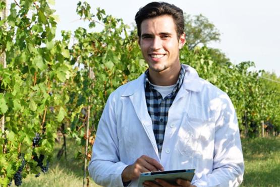 Mirando al frete: Joven con con sonrisa, viste una bata blanca y sostiene una tablet,mientras realiza labores de estudio en el campo. Al fondo un cultivo alto.